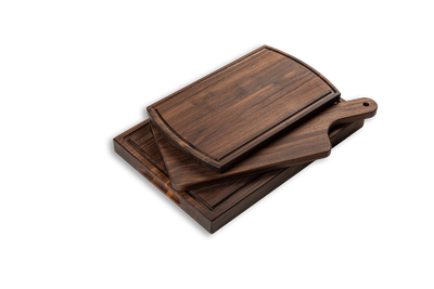 A set of three chop chop walnut wood cutting boards, varying in size, with a handle on the large serving board. They are displayed stacked, showcasing their different sizes and solid walnut wood grain.